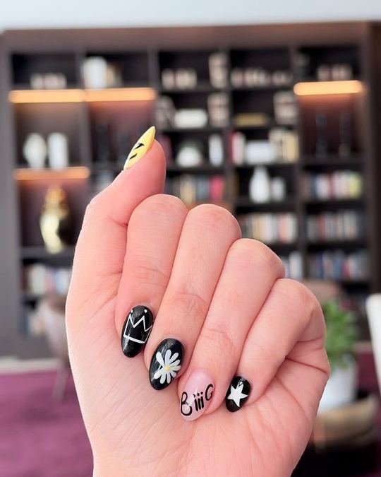 Short black nails with hand-painted white art: a crown, a daisy and a star, with one yellow accent nail